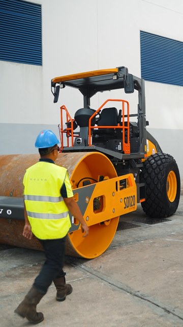 Video frame: a worker in a hi-vis vest walking past a Volvo SD120 roller
